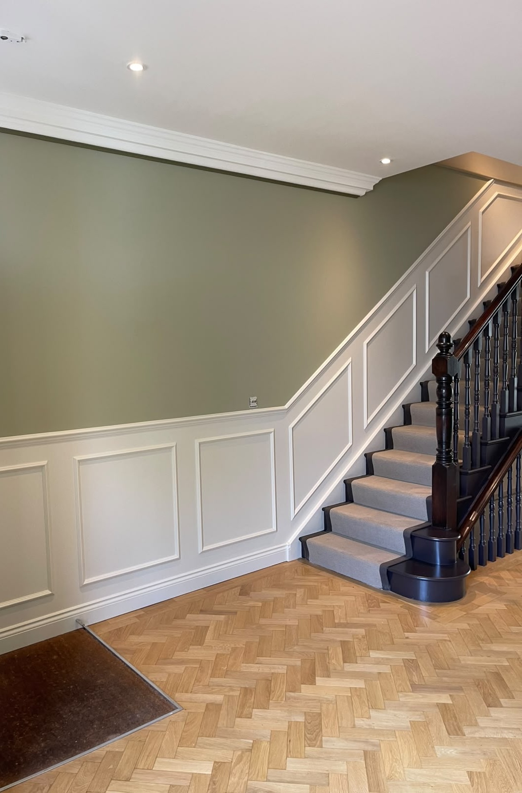 After: finished sage-green and white panelled staircase hallway with grey stair carpet, dark newel post and herringbone parquet floor