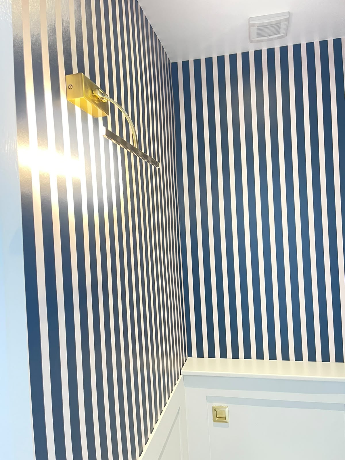 Cloakroom with blue and white striped wallpaper above painted wainscoting and brass picture light